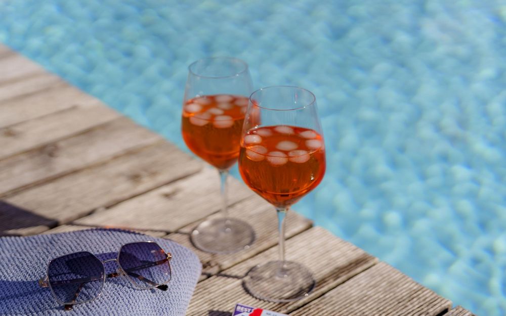 Zwei Gläser mit einem orangefarbenen Getränk und Eiswürfeln am Poolrand, daneben eine blaue Handtuch und eine Sonnenbrille.