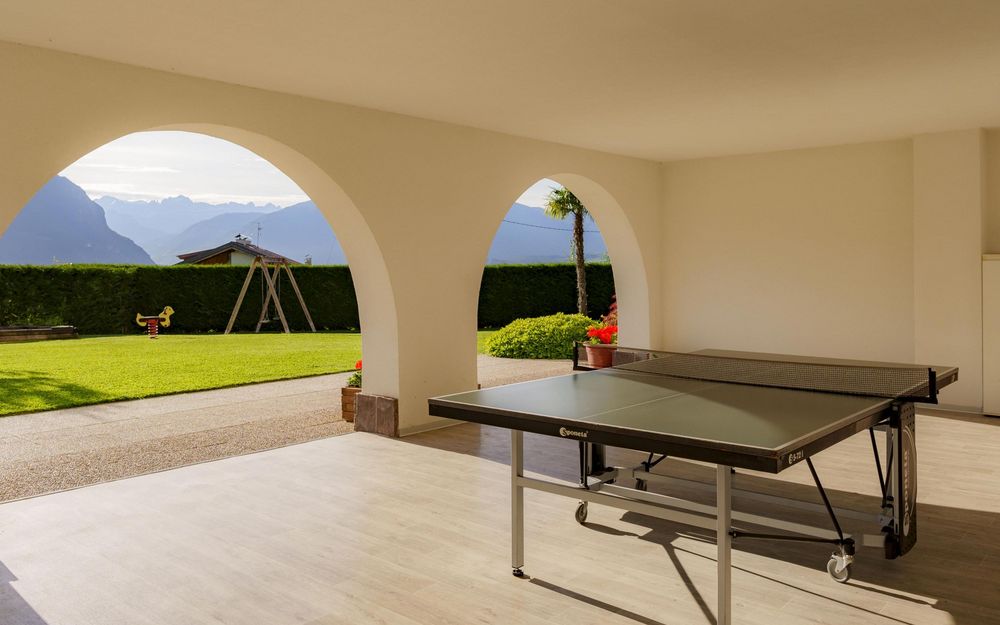 Pingpongtisch in einem überdachten Bereich mit Rundbögen und Blick auf einen Garten mit Spielplatz vor Bergen