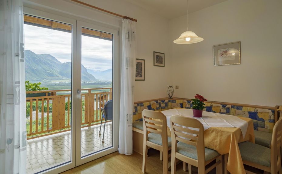 Sala da pranzo con angolo salotto, tavolo apparecchiato, sedie di legno, balcone con vista sulle montagne.