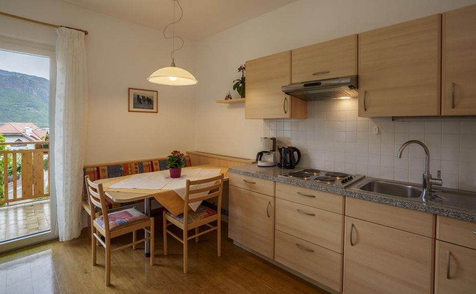 Cucina luminosa con mobili in legno chiaro, tavolo con sedie e piante, finestra con tenda bianca e vista montagna.