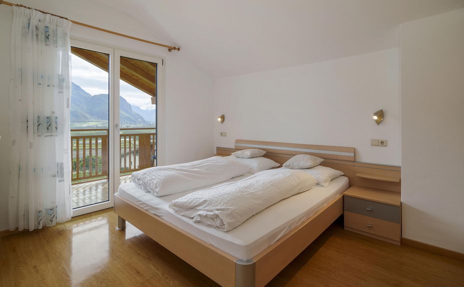 Minimalist bedroom with double bed, white bedding, wooden floor, and glass doors opening to a balcony with mountain view.