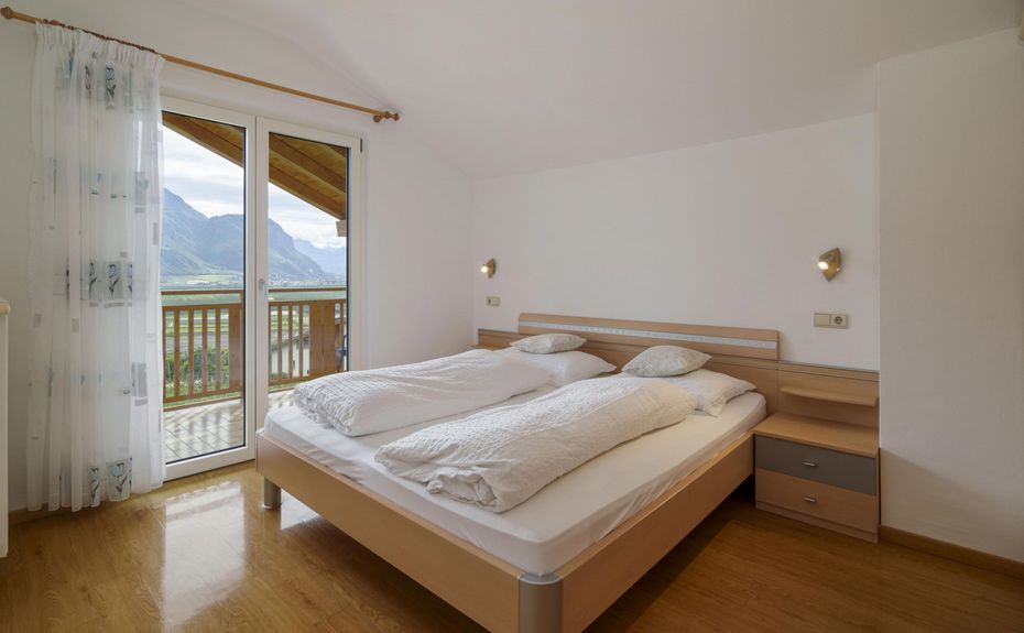 Camera da letto luminosa con letto matrimoniale, parquet, balcone con vista sulle montagne e tende bianche trasparenti.